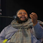 Michael Twitty, a Black man with a plaid scarf around his neck and also wearing a kippa seated in front of a microphone.
