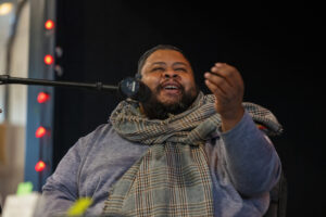 Michael Twitty, a Black man with a plaid scarf around his neck and also wearing a kippa seated in front of a microphone.