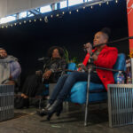 Three people seated on stage in front of microphones addressing and audience.