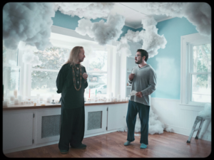 Movie still. Two men standing a light blue room with fluffy clouds surrounding them.